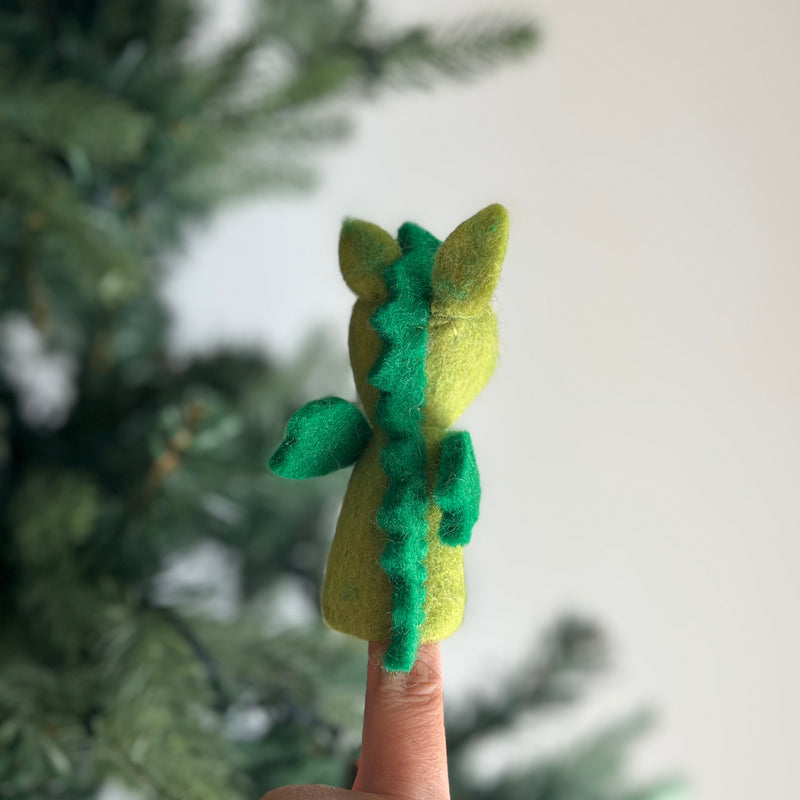 A handcrafted wool felt dragon finger puppet is worn on a finger, presented against a blurred green Christmas tree background. This vibrant, needle-felted dragon puppet has a light green body with darker green wings, tail, and a distinct spiky ridge running down its back. Its simple yet striking form makes it an engaging felt toy for kids.
