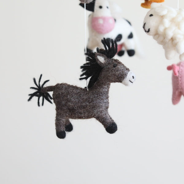 A close-up of a Farmland Animal Wool Felt Baby Mobile is set against a bright white background. In the foreground, a textured brown donkey, featuring black tufts for its mane and tail, hangs by a clear string. The mobile's cow and sheep figures are softly blurred in the background.
