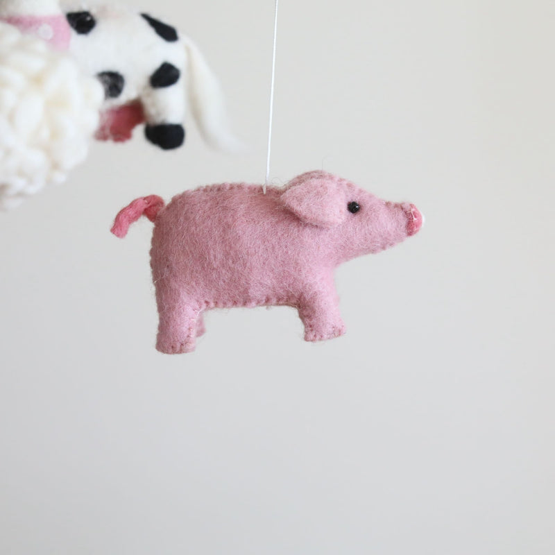 A pink felt pig element from the Farmland Animal felt baby mobile is shown in a close-up, hanging against a stark white background. Crafted from soft pink wool felt, the pig features a small black eye, a snout, and a curly tail with simple stitched details, highlighting its handmade charm. Blurry outlines of sheep and a cow are visible in the background, suggesting other elements of the whimsical mobile.