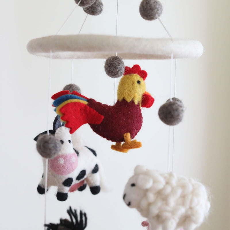 A wool felt Farmland Animal baby mobile is presented against a calm, off-white background. The whimsical mobile features a burgundy rooster with a yellow head and red comb in the center, flanked by a white cow with black spots and a white sheep, all suspended from a central ring. Gray wool felt balls are also attached.