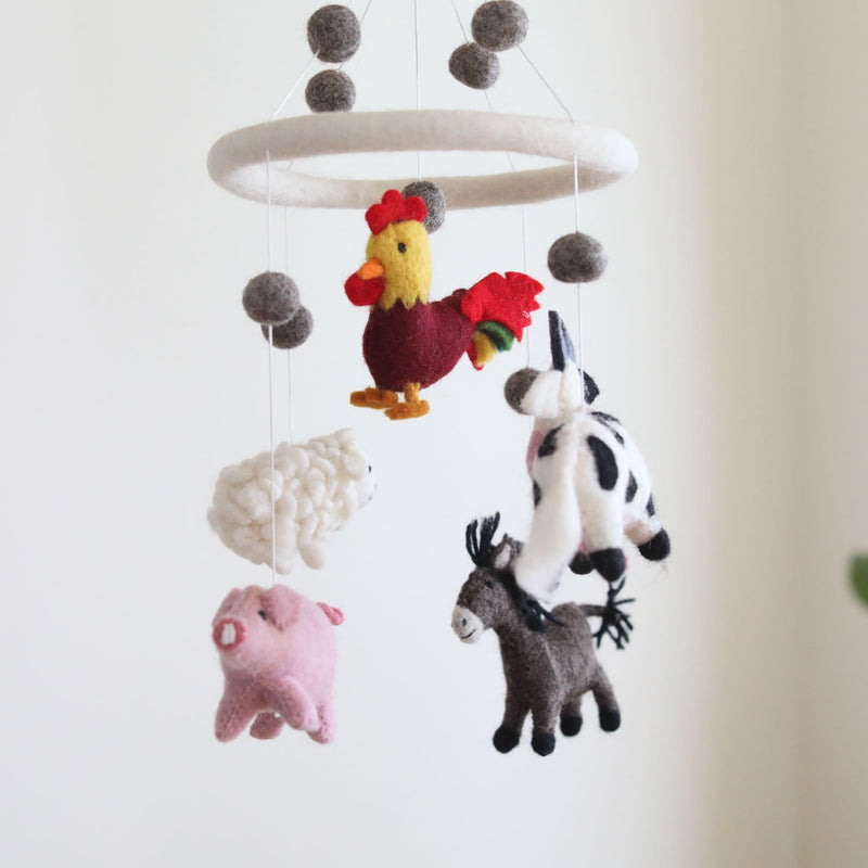 A Farmland Animal wool felt baby mobile hangs suspended against a light, neutral background, creating a calm and whimsical atmosphere. This handmade mobile features plush farm animals: a yellow rooster with red accents, a fluffy white sheep, a light pink pig, a black and white milk cow, and a gray donkey with a black mane. Gray felt balls and a white felt ring complete the mobile, adding balance and visual interest.