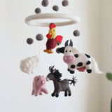 A farmland animal wool felt baby mobile is displayed against an off-white background. It features a white felt ring from which five colorful wool felt farm animals – a red and yellow rooster, a white and black spotted milk cow, a pink pig, a white sheep, and a brown donkey – are suspended by delicate threads. Grey felted balls hang above the animals, adding to the whimsical and comforting design of this nursery baby mobile.