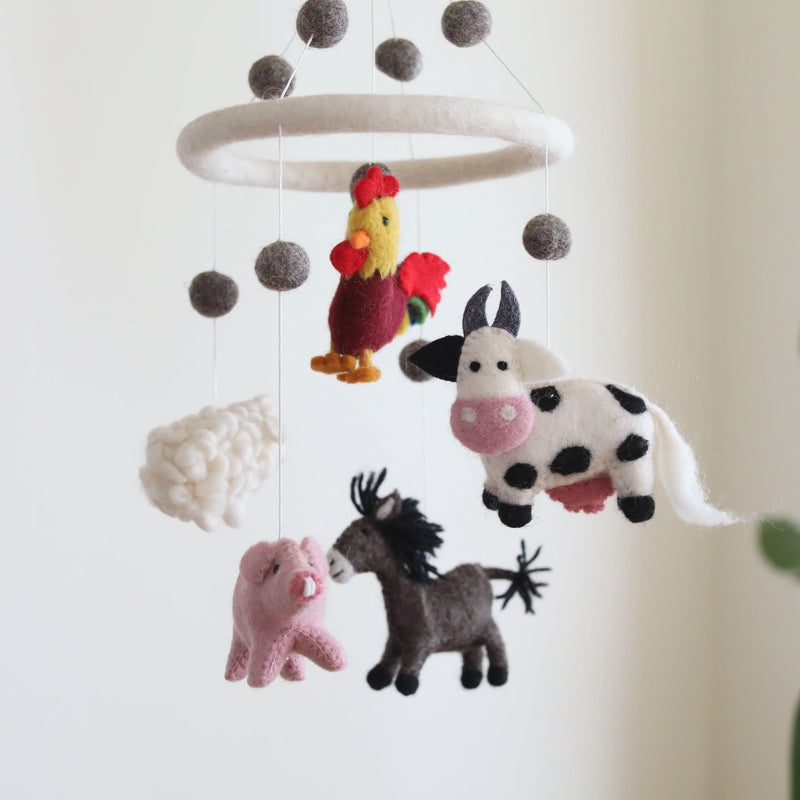 A farmland animal wool felt baby mobile is displayed against an off-white background. It features a white felt ring from which five colorful wool felt farm animals – a red and yellow rooster, a white and black spotted milk cow, a pink pig, a white sheep, and a brown donkey – are suspended by delicate threads. Grey felted balls hang above the animals, adding to the whimsical and comforting design of this nursery baby mobile.