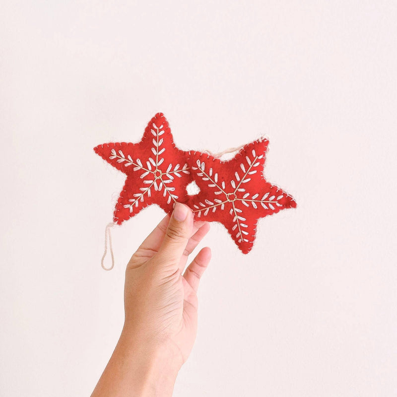 Two red felt star Christmas ornaments, part of a Snowflake Christmas Ornaments set, are delicately held by a female hand against a pale background. These handcrafted felt ornaments are made from red felt, featuring intricate white snowflake embroidery on both sides. The conjoined star ornaments include a white cotton string for hanging, adding festive elegance to any holiday decor.