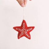 A red felt snowflake star Christmas ornament is delicately held by a female hand with polished nails against a bright, clean background. This handmade felt ornament features intricate white snowflake embroidery and hangs from a thin cotton string, emphasizing its handcrafted quality.