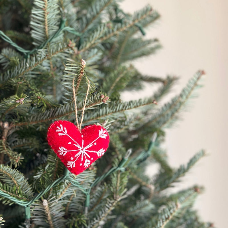 A bright red felt heart-shaped Christmas ornament, part of a set of snowflake Christmas ornaments, hangs by a jute string from a lush green Christmas tree branch. The handmade felt ornament features delicate white snowflake embroidery, contributing to a joyful, festive atmosphere.