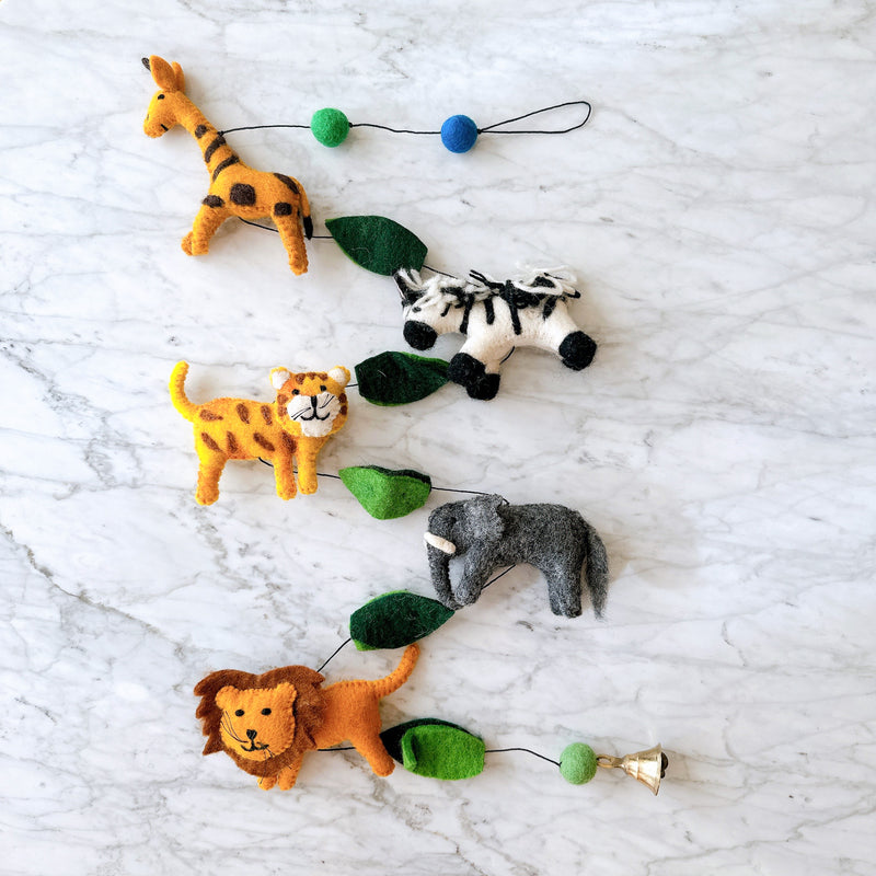A vibrant wool felt African safari animal garland is playfully displayed on a white marble surface. This handmade felt garland features five detailed 3D animals: a brown-spotted giraffe, an orange-striped tiger, a black and white striped zebra, a gray elephant, and a lion with a brown mane. Green felt leaves and blue and green felt balls separate the animals along a thin black string, ending with a small copper bell.