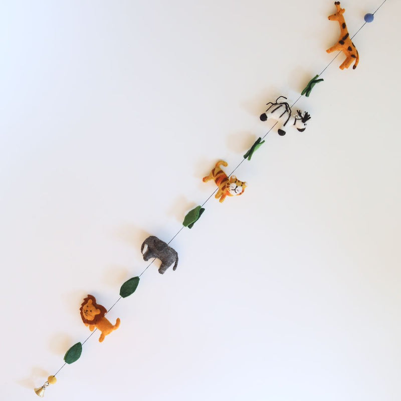 A cheerful wool felt African safari animal garland is displayed diagonally against a bright white background. This needle-felted, 3D felt garland features alternating animals on a thin black string. It showcases an orange lion, gray elephant, striped tiger, zebra, and spotted giraffe with a blue felt ball, ending with a copper bell.