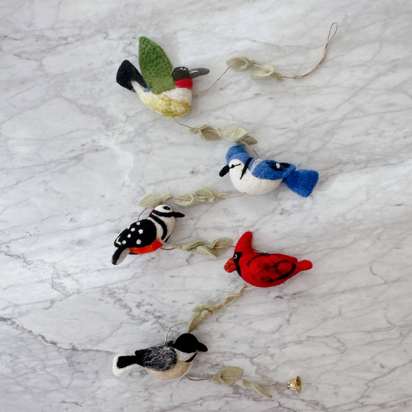 A vibrant felt American bird garland, featuring a green-winged hummingbird, a blue jay, a red cardinal, and a chickadee, is displayed diagonally on a light, marbled surface. The felt birds are connected by a wooden frame interspersed with felt leaves.