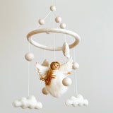 A felt angel baby mobile, made from wool felt, hangs from the ceiling against a white background. The central figure of an angel holding a halo is surrounded by clouds and stars, creating a soft and pastel color palette. The gentle and serene appearance evokes a sense of comfort and tranquility, perfect for a nursery or child's room.