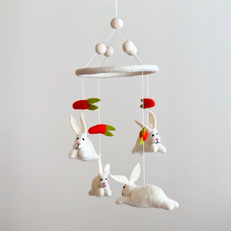 A felt baby mobile featuring bunnies and carrots is suspended against a plain white background. White felt balls at the top connect via strings to a felt ring. From the ring hang three fluffy white felt bunnies with pink noses and black eyes, alongside two vibrant red-orange felt carrots with green leafy tops.