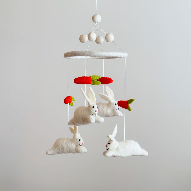 A charming felt baby mobile, featuring white bunnies and bright orange carrots, is suspended against a light gray background. The whimsical design showcases detailed bunnies with pink noses and stitched eyes, appearing to interact with carrots topped with green felt. Above the central felt ring, white felt balls and delicate strings support this playful and imaginative mobile, ideal for a nursery.