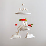 A whimsical felt baby mobile featuring bunnies and carrots is suspended against a simple off-white backdrop. This delightful nursery mobile has a circular felt frame, with three fluffy white felt bunnies and bright red felt carrots with green tops dangling on thin white strings. The bunnies appear to playfully chase the carrots at varying heights, creating a charming, playful design.