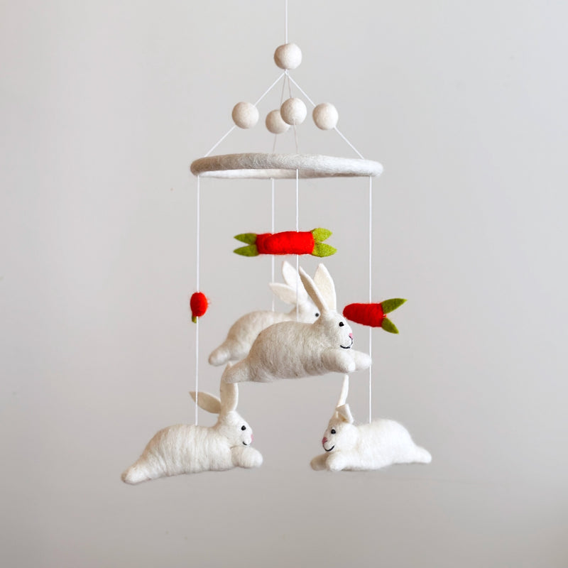 A whimsical felt baby mobile featuring bunnies and carrots is suspended against a simple off-white backdrop. This delightful nursery mobile has a circular felt frame, with three fluffy white felt bunnies and bright red felt carrots with green tops dangling on thin white strings. The bunnies appear to playfully chase the carrots at varying heights, creating a charming, playful design.