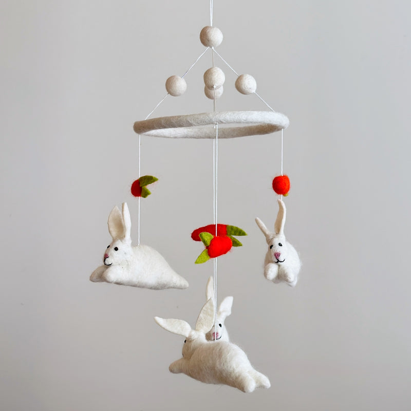 A playful felt baby mobile featuring bunnies and carrots floats gently against a muted light gray backdrop. Thin white strings suspend beige felt balls and a larger felt-covered ring. From this ring, three plush white felt bunnies in dynamic poses with black eyes and pink noses dangle alongside bright orange felt carrots with green tops. This charming and soft baby crib mobile creates a whimsical scene.