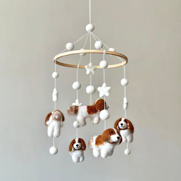 A whimsical wool felt baby mobile, featuring Cavalier King Charles spaniels, hangs suspended against a plain light gray backdrop. Its circular wooden frame supports dangling soft felt spaniels in white and brown, some standing and some sitting, interspersed with felt stars. The mobile's delicate design and charming white, beige, and brown colors create a dreamy, comforting nursery accent.