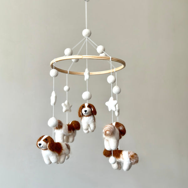 A wool felt Cavalier King Charles baby mobile is suspended against a plain light beige background, evoking a dreamy and gentle nursery ambiance. It features a wooden frame from which strings of white felt stars and four charming white and brown felt Cavalier King Charles spaniels hang at varying heights. A string with a loop is at the top for hanging.