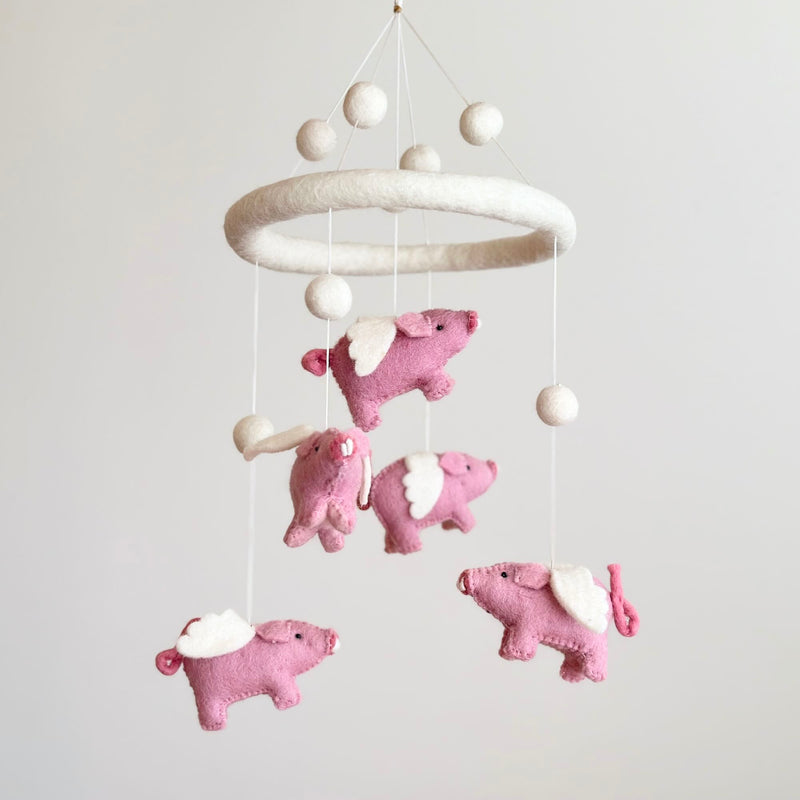 A handcrafted flying pig felt baby mobile is suspended against a plain, light background. Made from soft, natural wool felt, it features a circular felt hoop from which several pink pigs with white wings hang by thin white strings. The charming pigs have detailed curly tails and stitched facial features, creating a whimsical and comforting nursery accent.