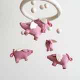 A whimsical wool felt baby mobile featuring pink flying pigs is suspended against a plain white background, creating a calm and airy mood. The mobile has a circular felt hoop suspending several soft, natural wool felt pink pigs with small white wings and curly tails. White wool felt balls are interspersed, adding a balanced, cloud-like aesthetic.