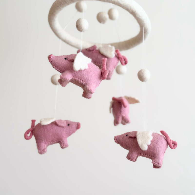 A whimsical wool felt baby mobile featuring pink flying pigs is suspended against a plain white background, creating a calm and airy mood. The mobile has a circular felt hoop suspending several soft, natural wool felt pink pigs with small white wings and curly tails. White wool felt balls are interspersed, adding a balanced, cloud-like aesthetic.