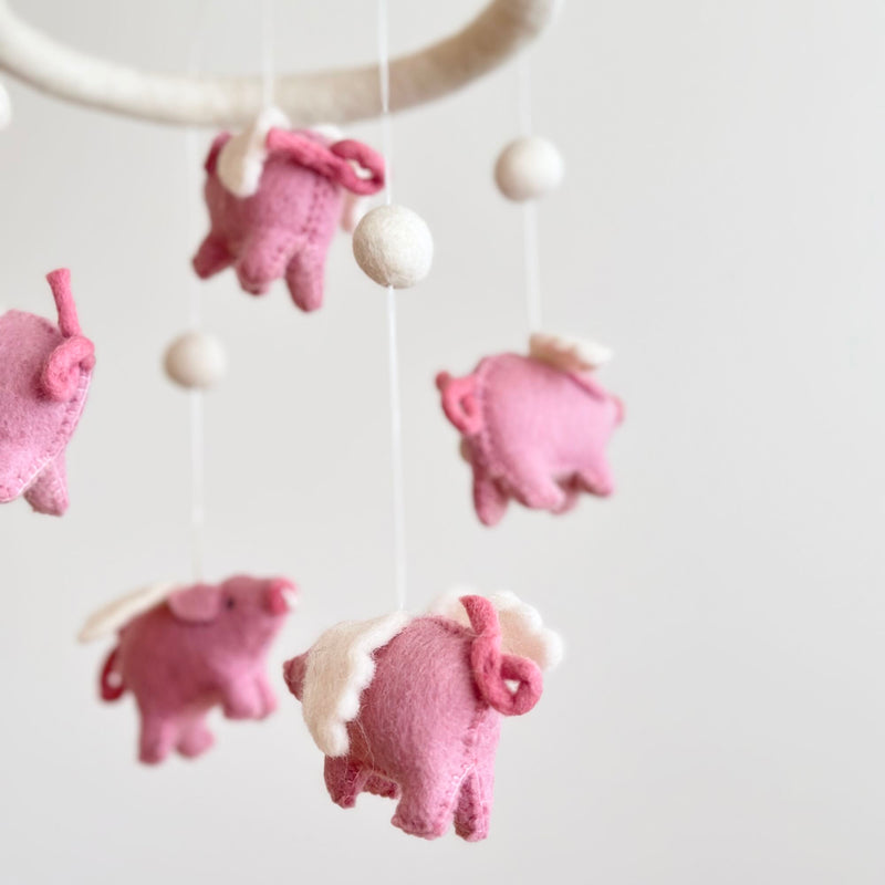A flying pig wool felt baby mobile is displayed against a soft white background. This handcrafted mobile features several pink wool felt pigs with white wings, suspended from a circular white wool felt hoop by delicate white strings. Small white wool felt balls are interspersed between the charming pigs, which each have a curled tail and detailed stitching.