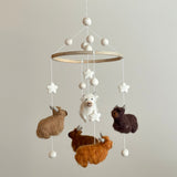 A Felt Baby Mobile featuring Highland Cows is displayed against a light gray background. The mobile has a wooden frame with soft cotton threads holding white felt balls and stars. Four felt Highland cows in white, caramel, brown, and chocolate dangle, showcasing intricate needlework, fluffy fur accents, and small felt horns. The mobile features a charming palette of white, beige, caramel, brown, and chocolate.