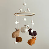 A felt baby mobile featuring Highland cows is displayed against a neutral background. The mobile features a wooden ring frame, suspended by white cotton threads, with white felted balls at the top. Four felt Highland cows in caramel, white, chocolate, and brown dangle from the ring, accompanied by small white stars and additional felted white balls. The cows have intricate needlework and fluffy fur accents, creating a playful and calming nursery mobile.