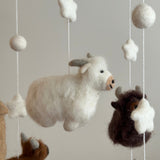 A felt Highland Cow baby mobile is suspended from a wooden frame against a plain, light gray background. It features felted white and brown Highland cows with fluffy fur accents, accompanied by white stars and spherical balls. The intricate needlework adds a playful and comforting touch.