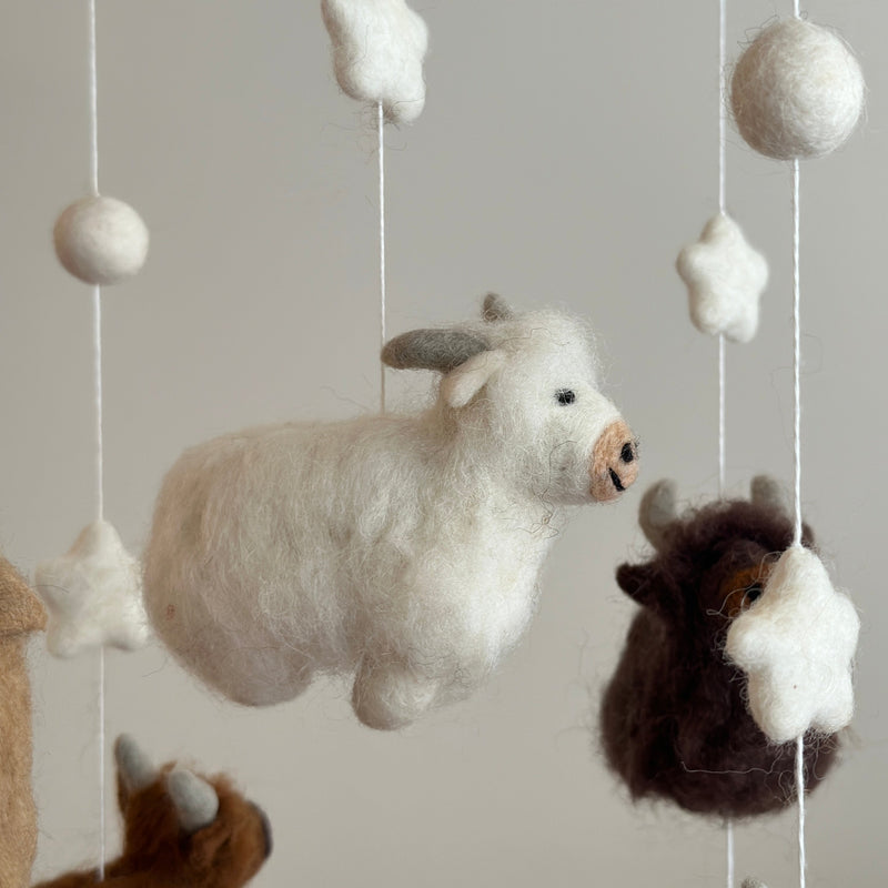 A felt Highland Cow baby mobile is suspended from a wooden frame against a plain, light gray background. It features felted white and brown Highland cows with fluffy fur accents, accompanied by white stars and spherical balls. The intricate needlework adds a playful and comforting touch.