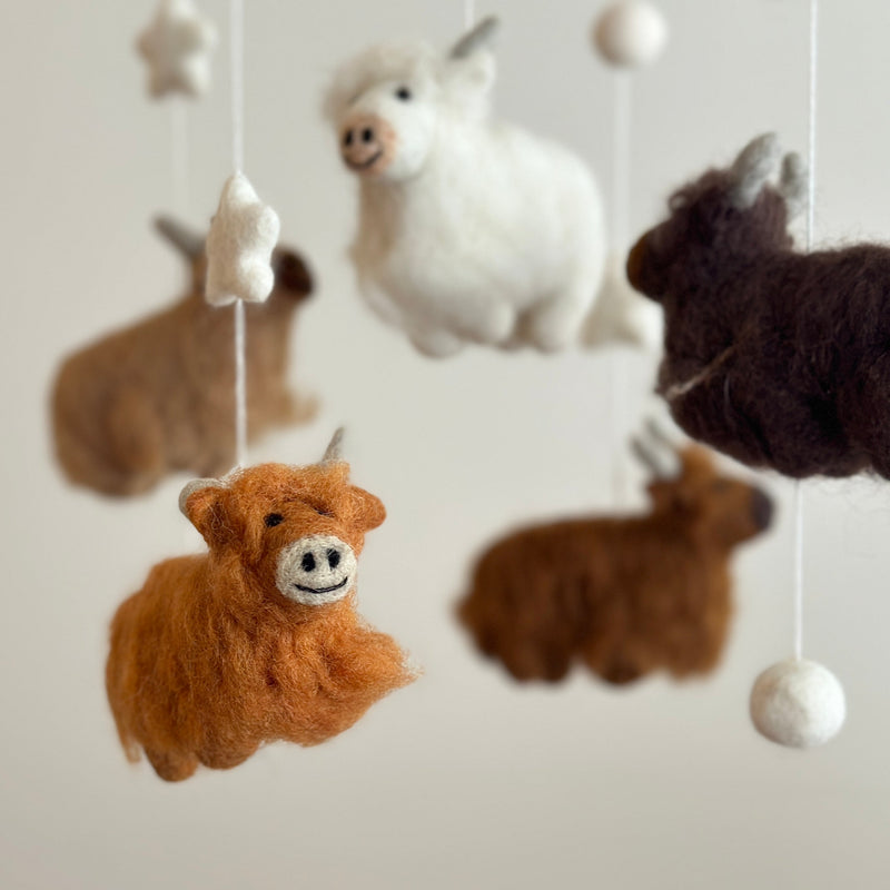 A delightful felt baby mobile featuring Highland cows is captured in a close-up studio shot against a soft white background. A charming caramel-colored felt Highland cow with fluffy fur accents and a sweet stitched smile hangs from a wooden frame. In the blurred background, additional white, beige, and chocolate felt cows, along with white felt stars and balls, create a playful scene.