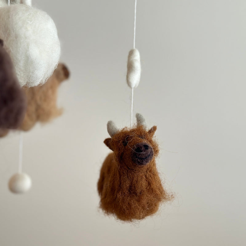 A felt Highland Cow baby mobile hangs centrally against an off-white backdrop. It features a wooden frame suspending various felt pieces in a warm palette of white, beige, caramel, brown, and chocolate. A brown felt Highland Cow with fluffy fur and small white horns is prominent in the foreground, with a white puff ball and a caramel Highland Cow above it.