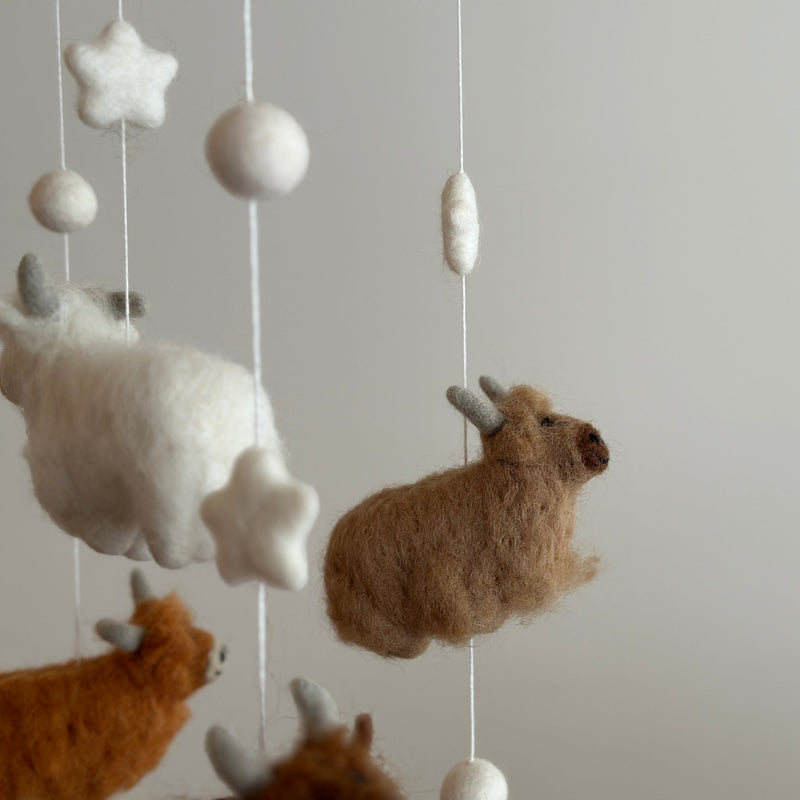 A wool felt baby mobile, featuring a highland cow, is displayed against a neutral background. A fluffy, caramel-colored wool felt highland cow with intricate needlework hangs from a wooden frame, accompanied by white felt stars and round balls. The mobile's gentle white, beige, caramel, brown, and chocolate colors create a whimsical and comforting nursery decoration.
