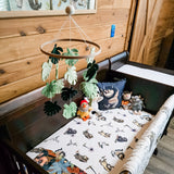 A cozy nursery setting featuring a wooden crib at the foreground, topped by our wool felt baby mobile - Monstera Serenity. The mobile showcases various monstera leaf designs in three different green shades, suspended above the crib. A stuffed animal and small toy are placed in front of the crib, while a wooden wall with a framed picture creates a warm and inviting ambiance.