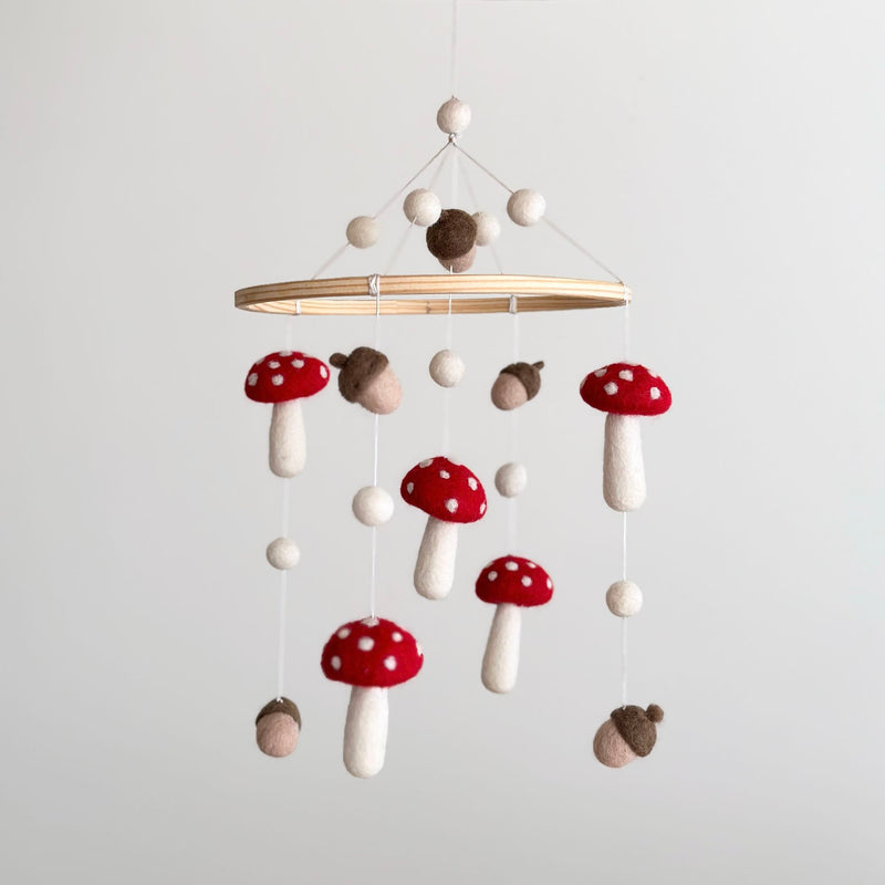 A whimsical wool felt baby mobile featuring mushrooms and acorns is gracefully suspended against a serene, light gray backdrop. Handcrafted red-capped mushrooms with white dots and brown acorns are delicately hung from a smooth, light wood hoop. Small, off-white spherical felt balls connect the natural wool felt figures, creating a playful yet calming design ideal for a nursery.