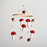 A felt baby mobile featuring mushrooms and acorns is suspended in mid-air against a light gray background. It showcases a smooth wooden hoop from which handcrafted wool felt shapes hang on delicate strings. The warm, earthy design includes bright red mushrooms with white spots and white stems, brown and tan acorns, and small white felt balls above the hoop and between the hanging elements. This unique baby mobile evokes a sense of woodland magic.