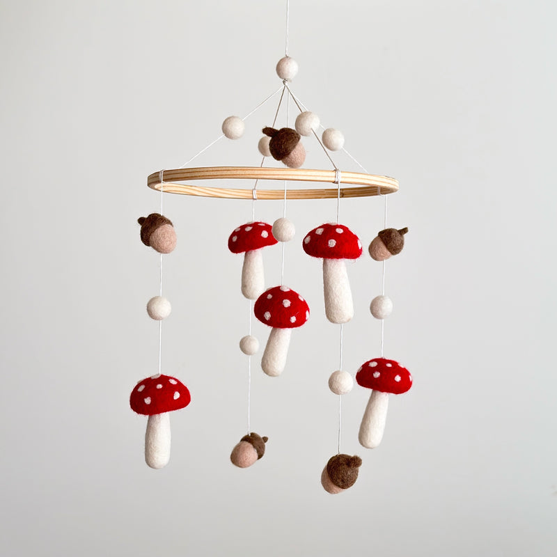 A felt baby mobile featuring mushrooms and acorns is suspended in mid-air against a light gray background. It showcases a smooth wooden hoop from which handcrafted wool felt shapes hang on delicate strings. The warm, earthy design includes bright red mushrooms with white spots and white stems, brown and tan acorns, and small white felt balls above the hoop and between the hanging elements. This unique baby mobile evokes a sense of woodland magic.