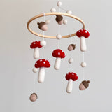 A wool felt baby mobile, featuring mushrooms and acorns, is presented indoors against a plain, light gray background. The mobile showcases handcrafted 100% wool felt mushrooms with red caps and white dots, alternating with earthy brown and light tan felt acorns. These pieces hang from a smooth, natural light-colored wooden hoop by thin white strings, with small white felt balls at the top, creating a whimsical woodland design.