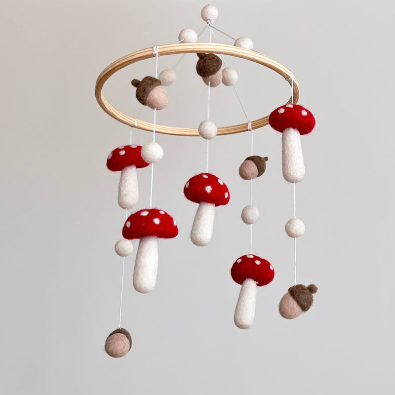 A wool felt baby mobile, featuring mushrooms and acorns, is presented indoors against a plain, light gray background. The mobile showcases handcrafted 100% wool felt mushrooms with red caps and white dots, alternating with earthy brown and light tan felt acorns. These pieces hang from a smooth, natural light-colored wooden hoop by thin white strings, with small white felt balls at the top, creating a whimsical woodland design.