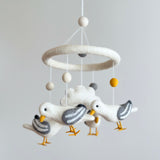 A felt seagull baby mobile hangs in a brightly lit space against a plain white background. It features four white felt seagulls with outstretched wings, grey wing stripes, and yellow beaks and feet, circling a central white felt cloud. Above the cloud, white, grey, and yellow felt balls hang from a white felt ring, with a white felt ball at the very top.