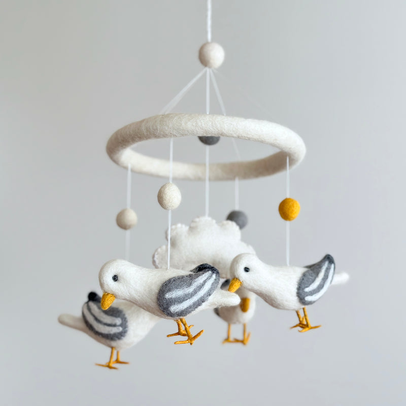 A felt seagull baby mobile hangs in a brightly lit space against a plain white background. It features four white felt seagulls with outstretched wings, grey wing stripes, and yellow beaks and feet, circling a central white felt cloud. Above the cloud, white, grey, and yellow felt balls hang from a white felt ring, with a white felt ball at the very top.
