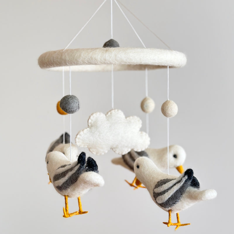 A felt baby mobile featuring four charming seagulls is presented against a light gray wall. Each handcrafted seagull has outstretched wings, with white bodies, gray striped wings, and yellow beaks and feet. The mobile's soft, dreamy aesthetic creates a calming atmosphere, perfect for a nursery.