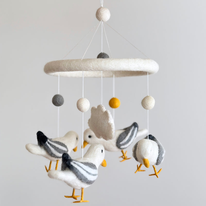 A felt seagull baby mobile is displayed against a plain white background. It features four handcrafted felt seagulls with outstretched wings, predominantly white with grey striped wings and bright yellow beaks and feet. They are suspended from a central ring, above which are white, grey, and yellow felt balls attached by white strings, creating a soothing design.