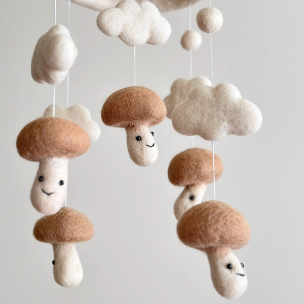 A white felt baby mobile, featuring five mushroom-shaped decorative pieces in shades of beige and white, suspended from the ceiling. Two of the mushrooms have smiley faces drawn on them, adding a playful touch. The overall design is whimsical and nature-themed, perfect for a nursery.
