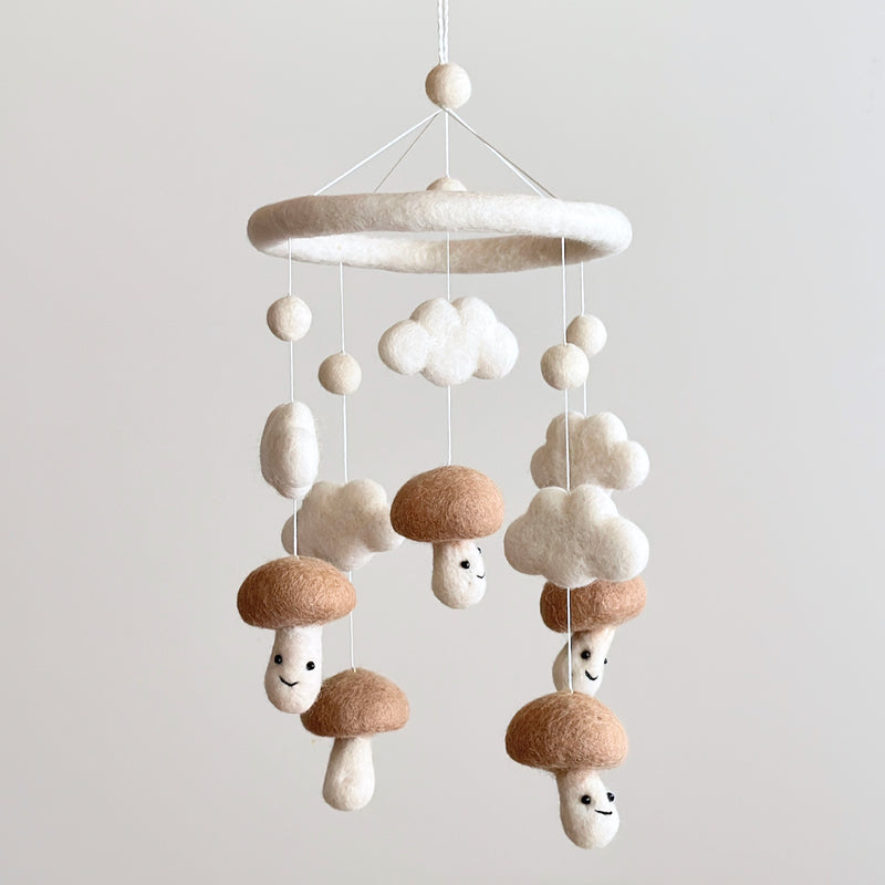A baby mobile made of wool felt, featuring five mushroom-shaped designs with smiling faces in beige color, mounted on a circular white frame resembling a cloud shape. The design is playful yet serene, reminiscent of childhood and nature-inspired themes.