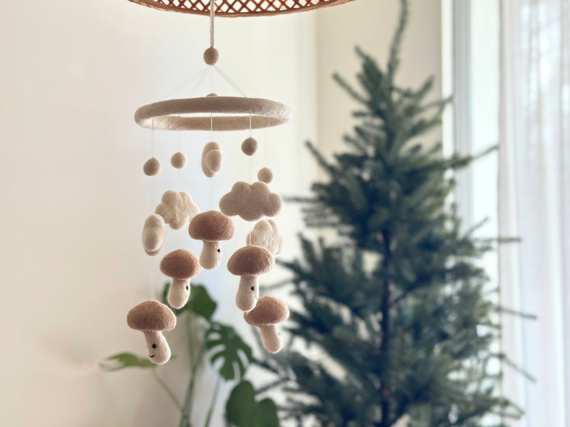 A cozy indoor setting featuring a felt baby mobile hanging from the ceiling, showcasing a white wool felt frame with five mushroom-shaped elements each bearing a smiling face. The background includes a Christmas tree adorned with ornaments and lights, suggesting a festive atmosphere.