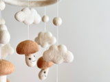 A whimsical felt baby mobile, featuring five smiley beige mushrooms on a white wool felt frame, suspended from a string. The mushrooms vary in size and shape, with some having smiling face designs.