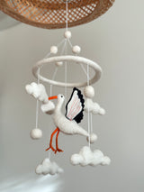 A felt baby mobile, featuring a stork design, hangs from a string suspended from a circular wooden frame. The mobile's soft pastel colors include shades of blue, pink, and white, with clouds, birds, and stars surrounding the central figure. Made from wool felt, this handcrafted nursery decor is a serene and whimsical addition to any child's bedroom.