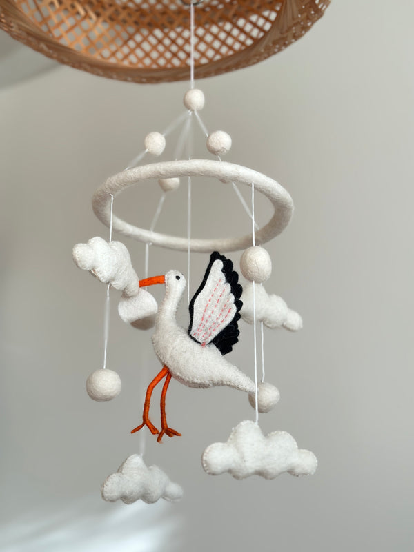 A felt baby mobile, featuring a stork design, hangs from a string suspended from a circular wooden frame. The mobile's soft pastel colors include shades of blue, pink, and white, with clouds, birds, and stars surrounding the central figure. Made from wool felt, this handcrafted nursery decor is a serene and whimsical addition to any child's bedroom.