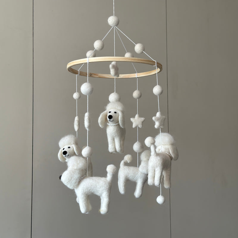 A white poodle wool felt baby mobile hangs against a plain gray wall, creating a soft, whimsical atmosphere. The mobile features a circular wooden frame from which several white, wool-felt poodles and a felted wool star are suspended by thin white strings. The delicate, handmade figures contribute to a clean and simple aesthetic.