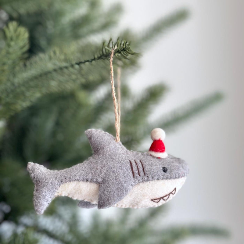 A wool felt baby shark ornament, complete with a red Santa hat, dangles from a Christmas tree branch. This charming felt baby shark ornament features a light gray upper body, a contrasting white lower section with stitched teeth, and a fluffy white pompom on its hat. A jute string allows it to hang playfully, adding a unique touch to festive decor.
